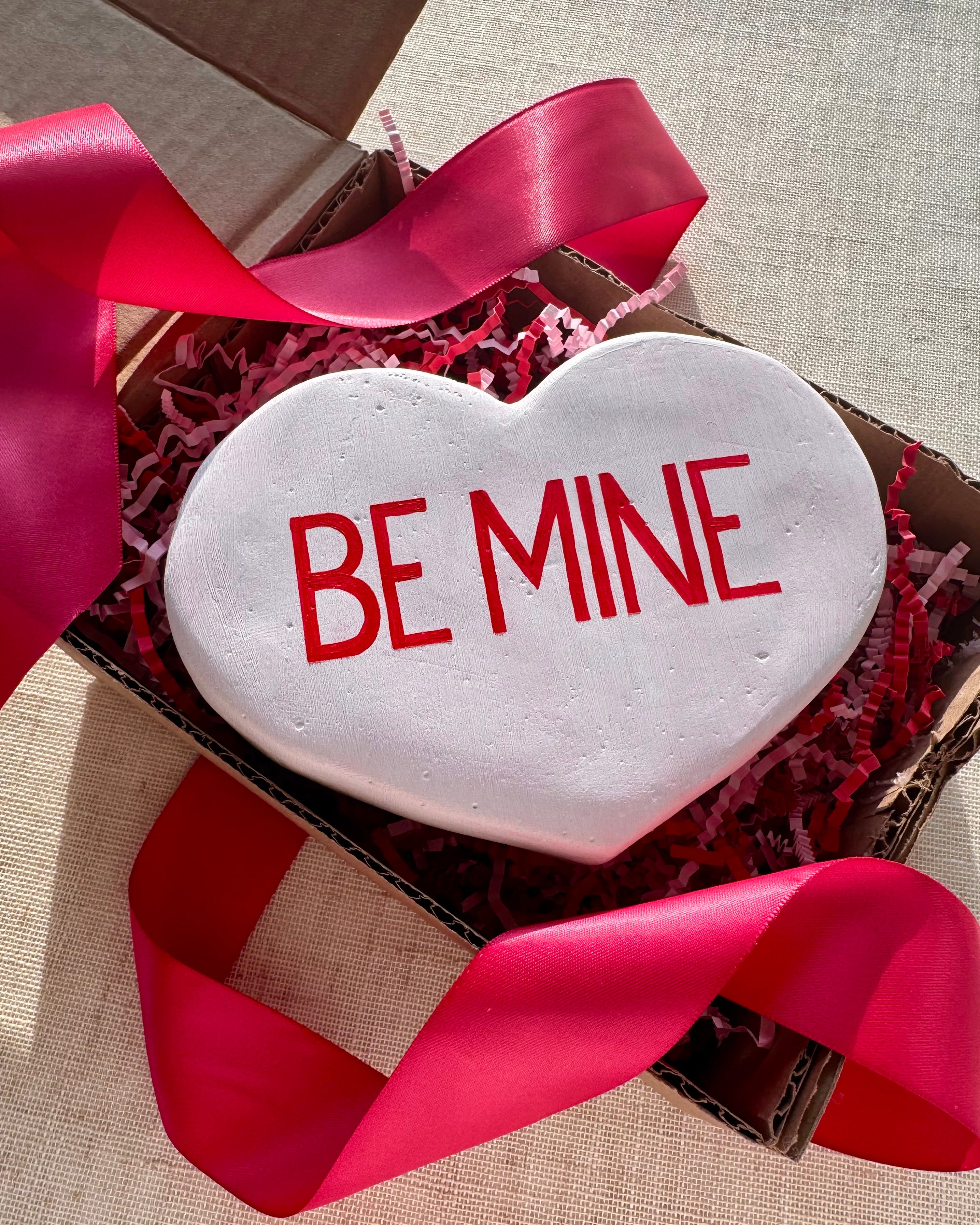 White heart-shaped sculpture with 'BE MINE' text in a box with red ribbon