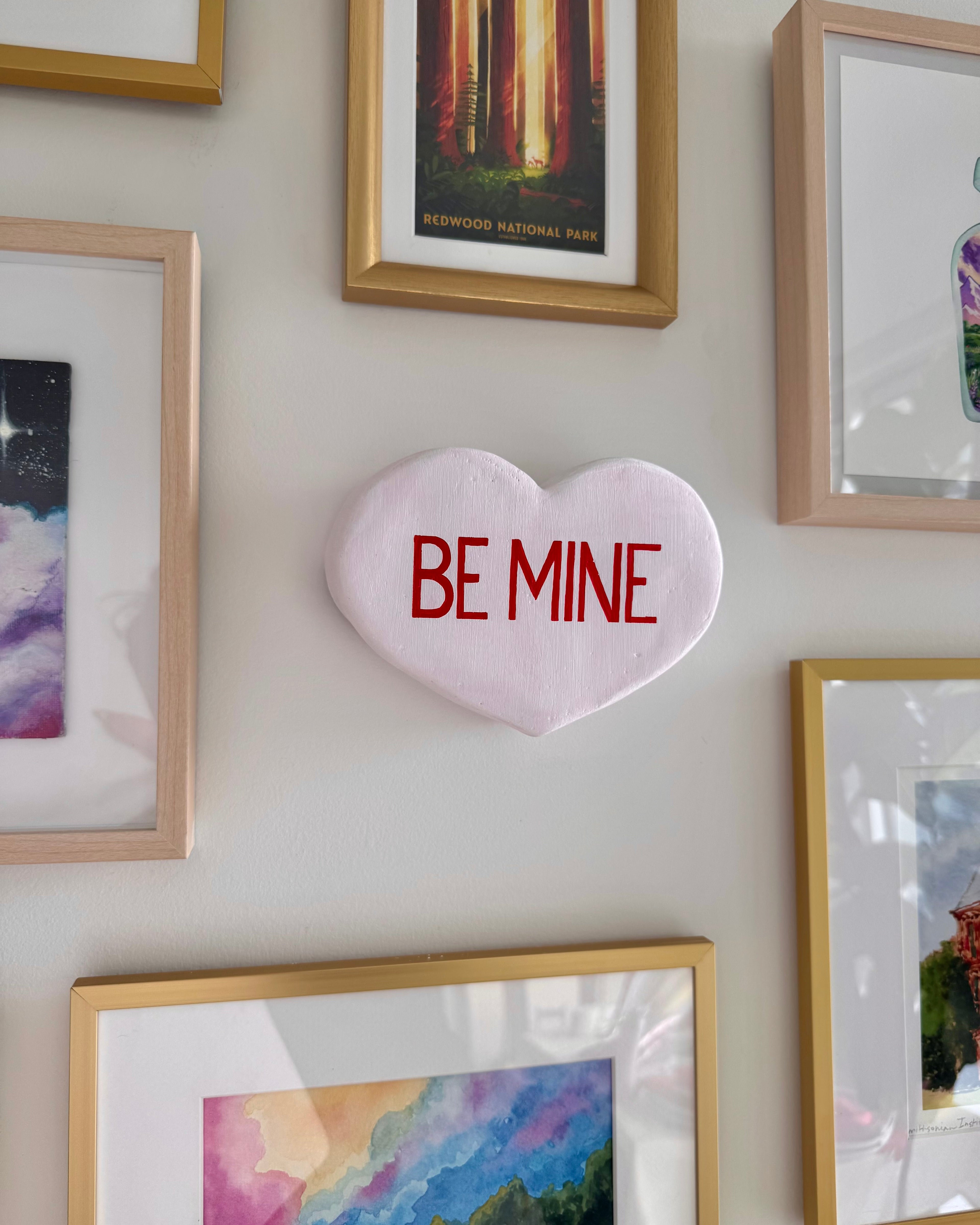 White heart-shaped scultpure with 'BE MINE' text on a wall with framed pictures