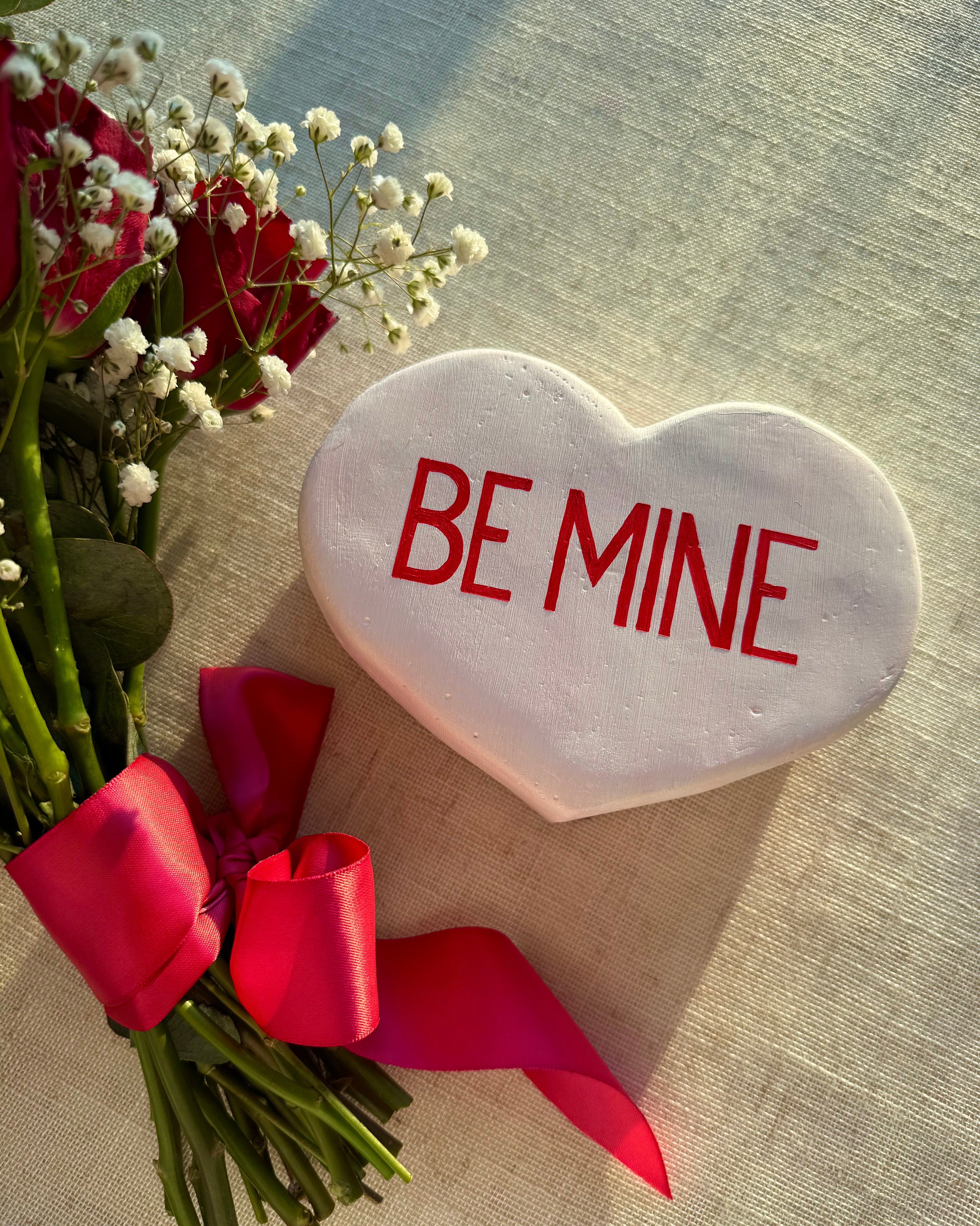 White heart-shaped sculpture with 'BE MINE' text surrounded by red roses and white flowers on a textured surface.
