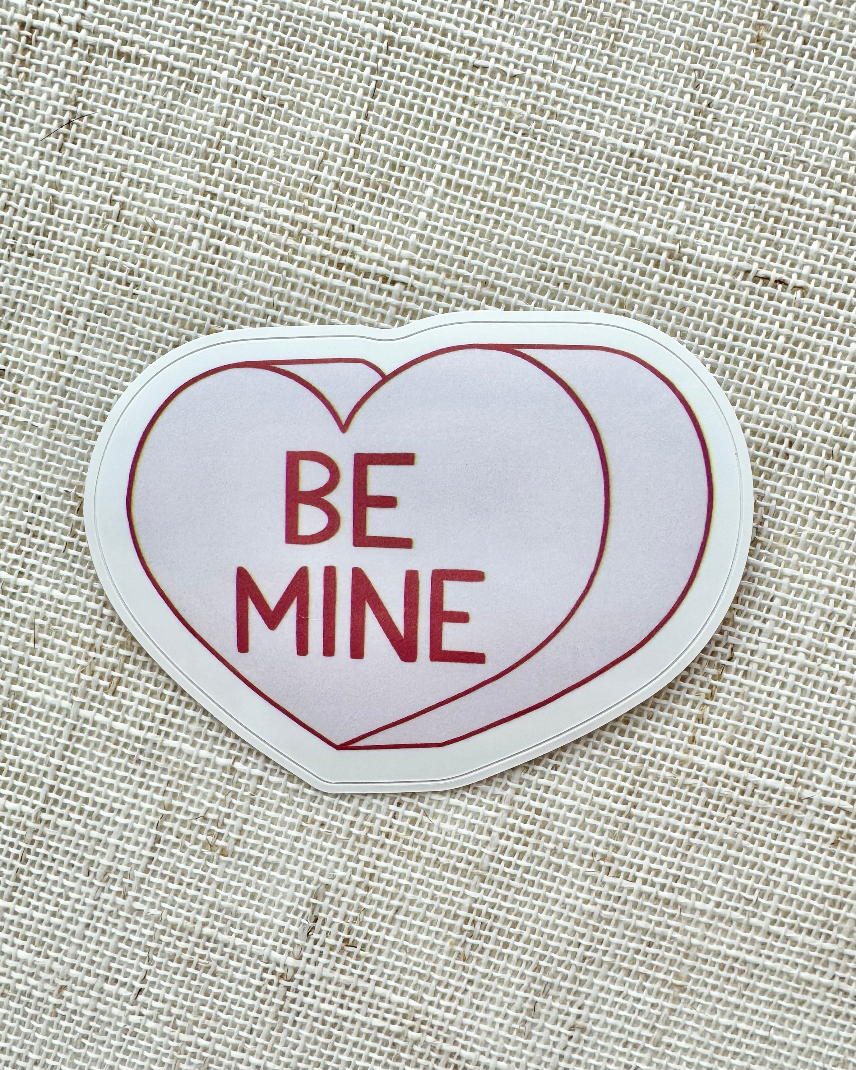 Heart-shaped sticker with 'BE MINE' text on a textured fabric background