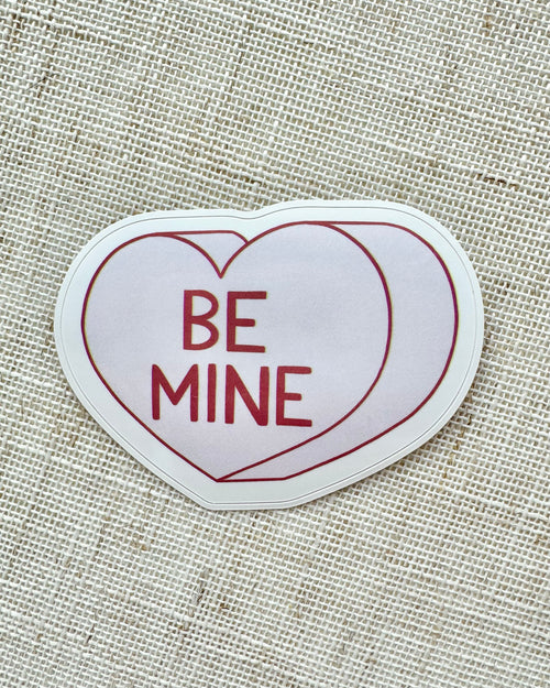 Heart-shaped sticker with 'BE MINE' text on a textured fabric background