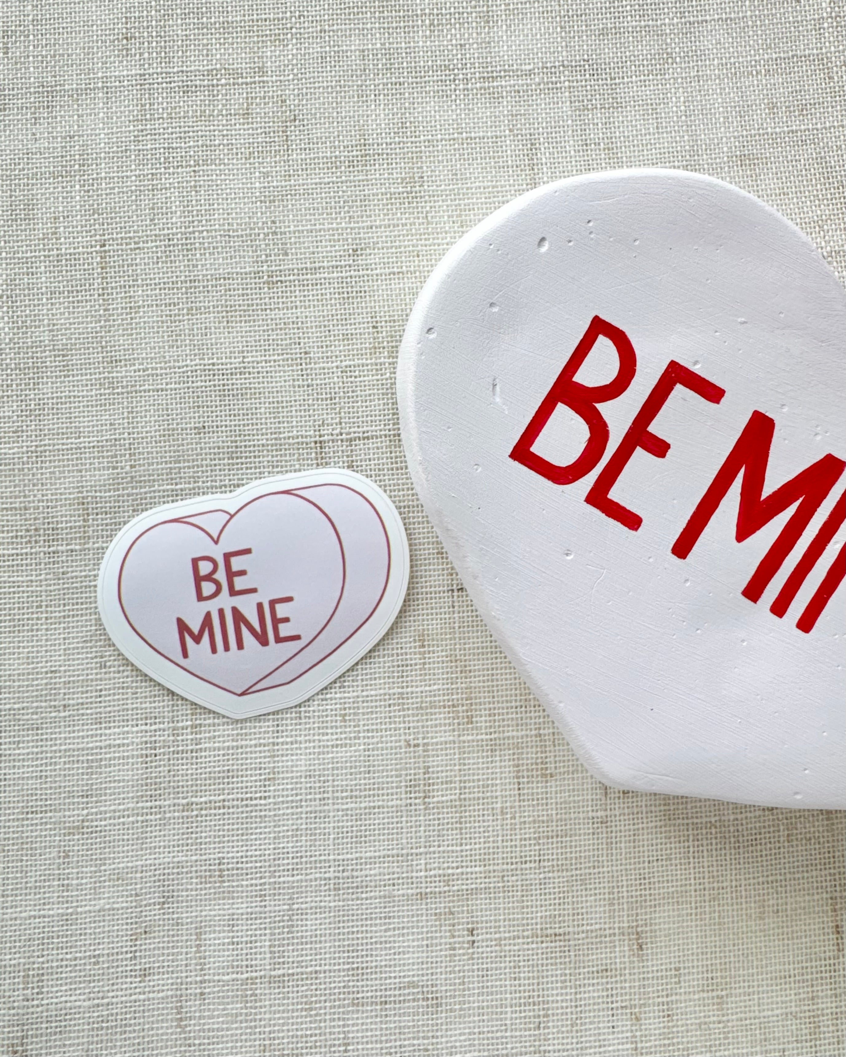 Heart-shaped sticker with 'BE MINE' text next to a heart-shaped sculpture with 'BE MINE' text on a textured fabric background