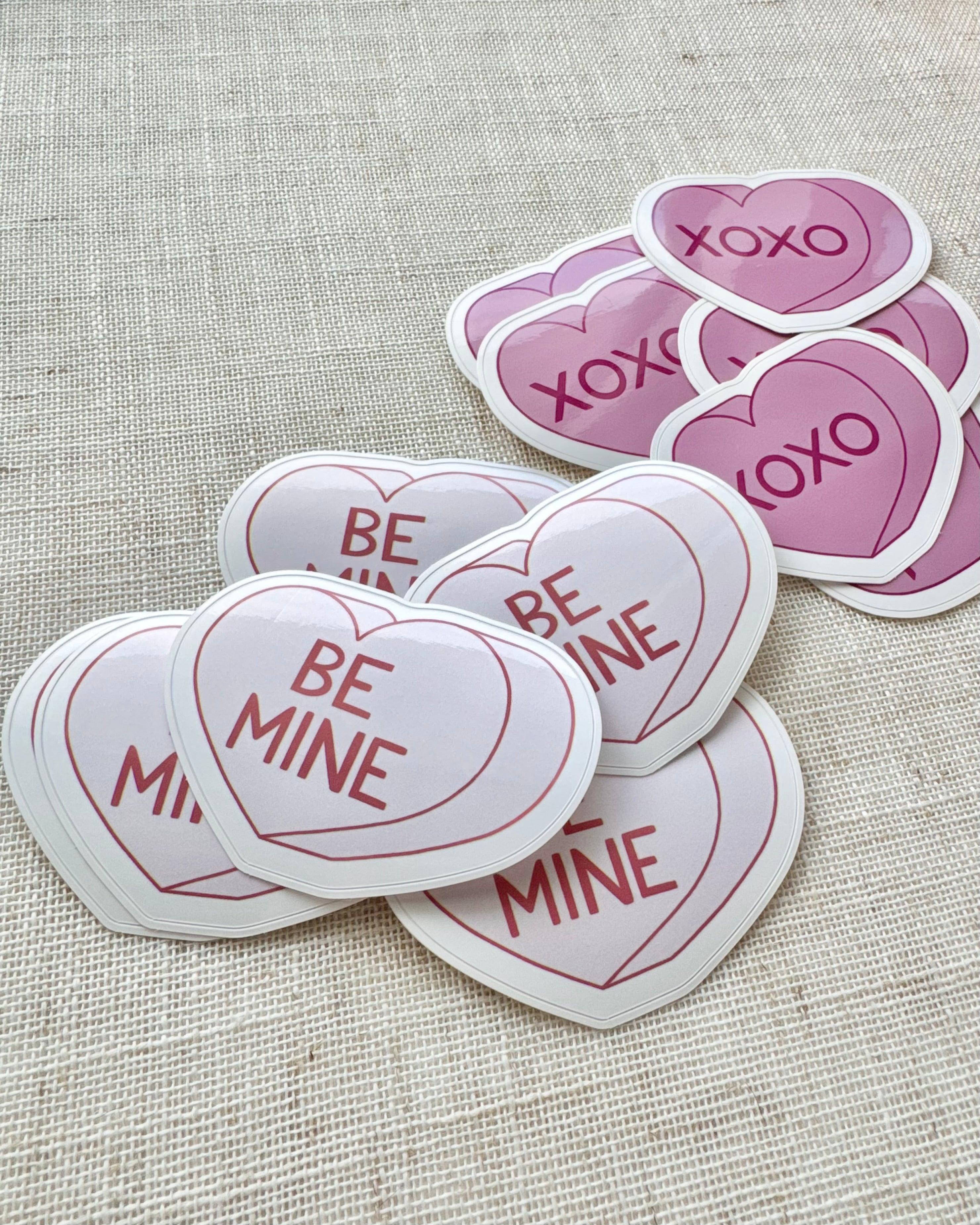 Heart-shaped stickers with 'Be Mine' and 'XOXO' text on a textured fabric background
