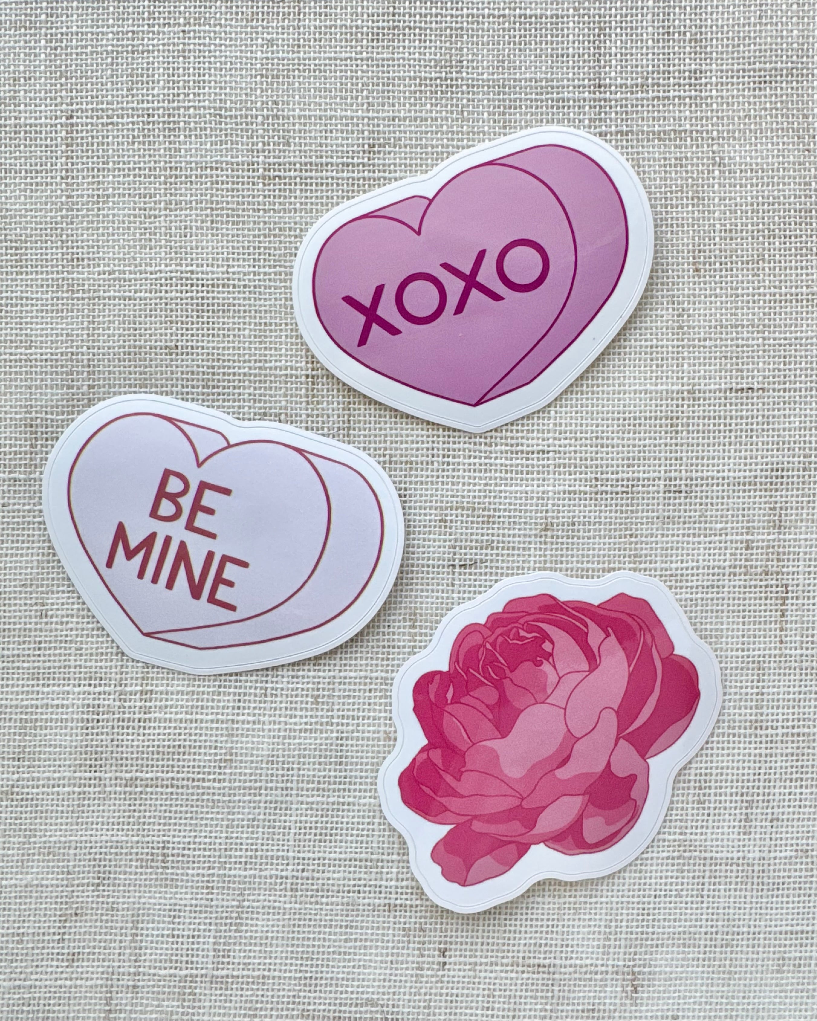 Three  stickers, two heart-shaped with 'XOXO', 'Be Mine', and a pink peony design on a textured fabric background.