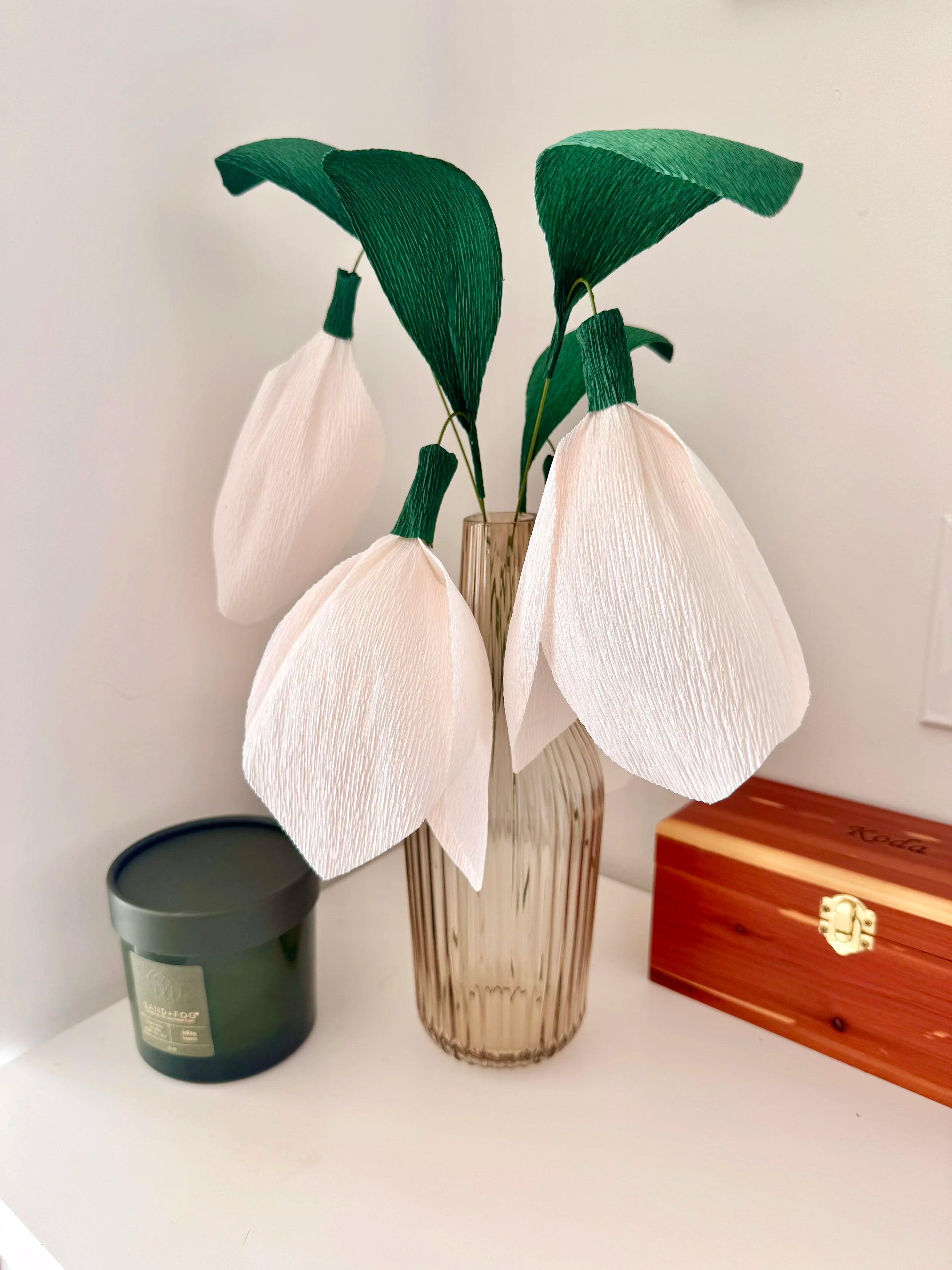 Decorative snowdrops with green leaves in a vase on a shelf.