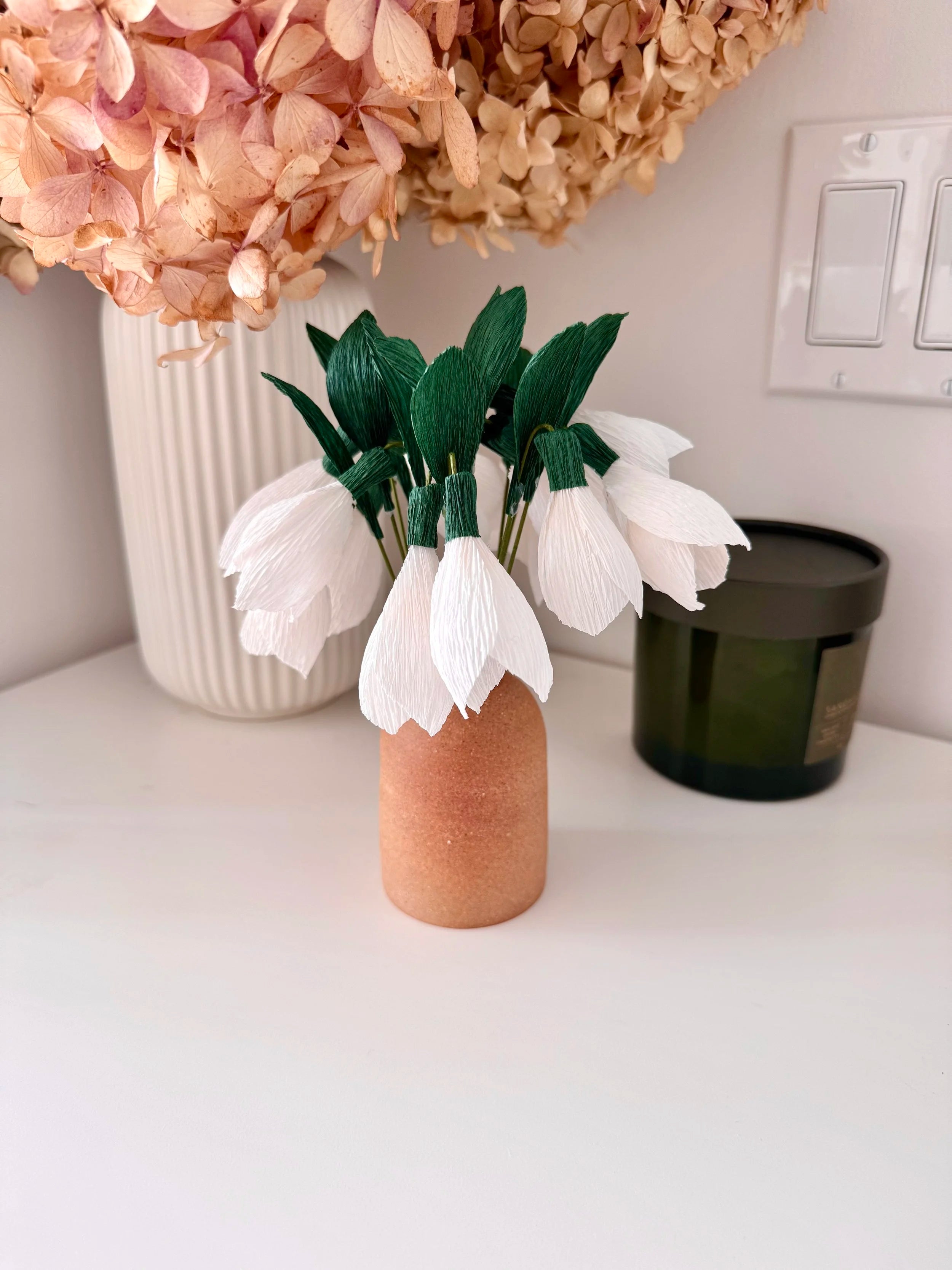 Small terracotta vase with white snowdrops on a white surface, with a larger bouquet of flowers in the background.