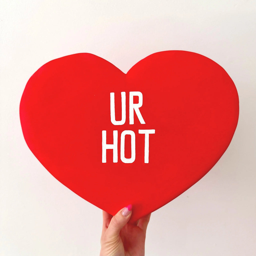 Red heart-shaped sculpture with 'UR HOT' text held by a hand on a light background