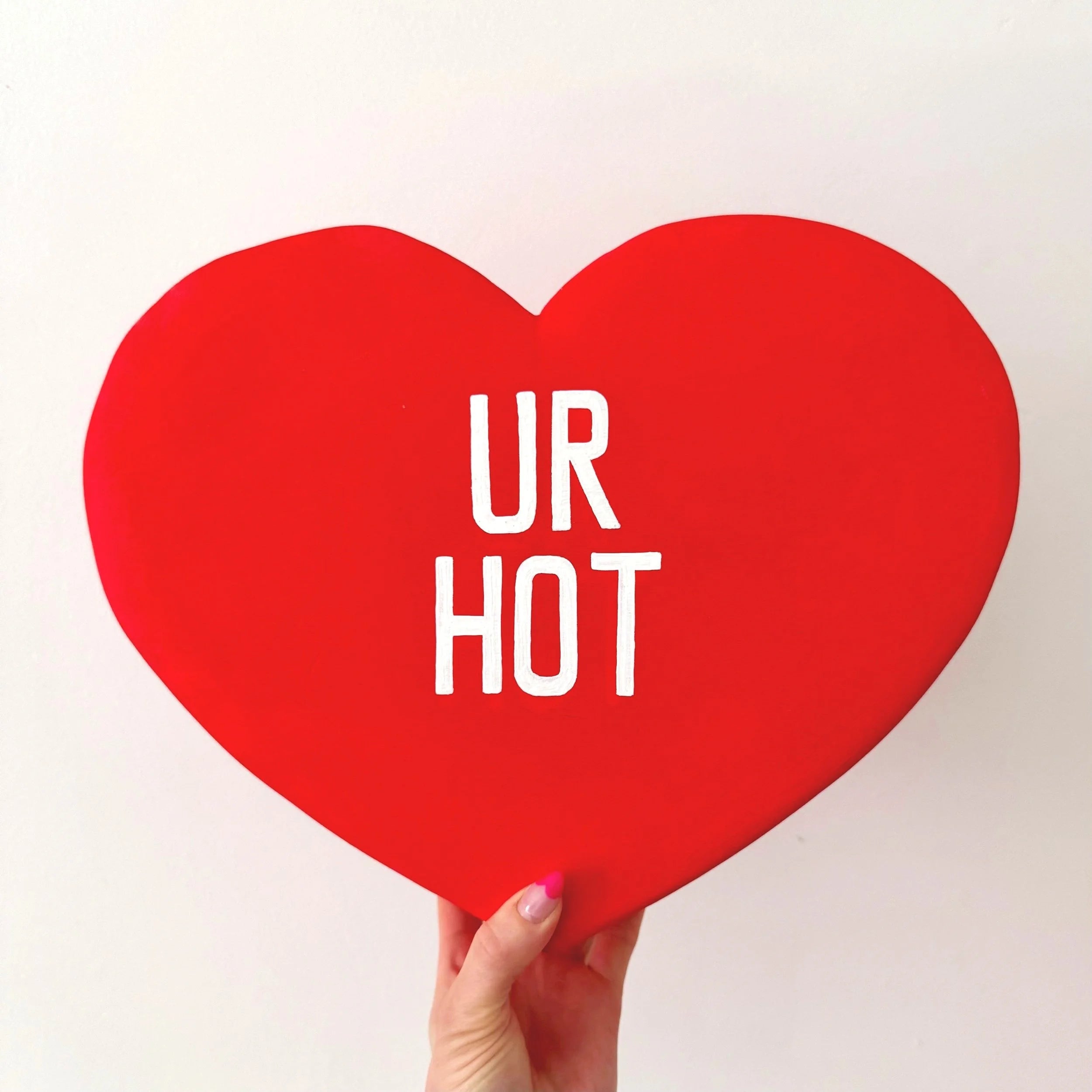 Red heart-shaped sculpture with 'UR HOT' text held by a hand on a light background