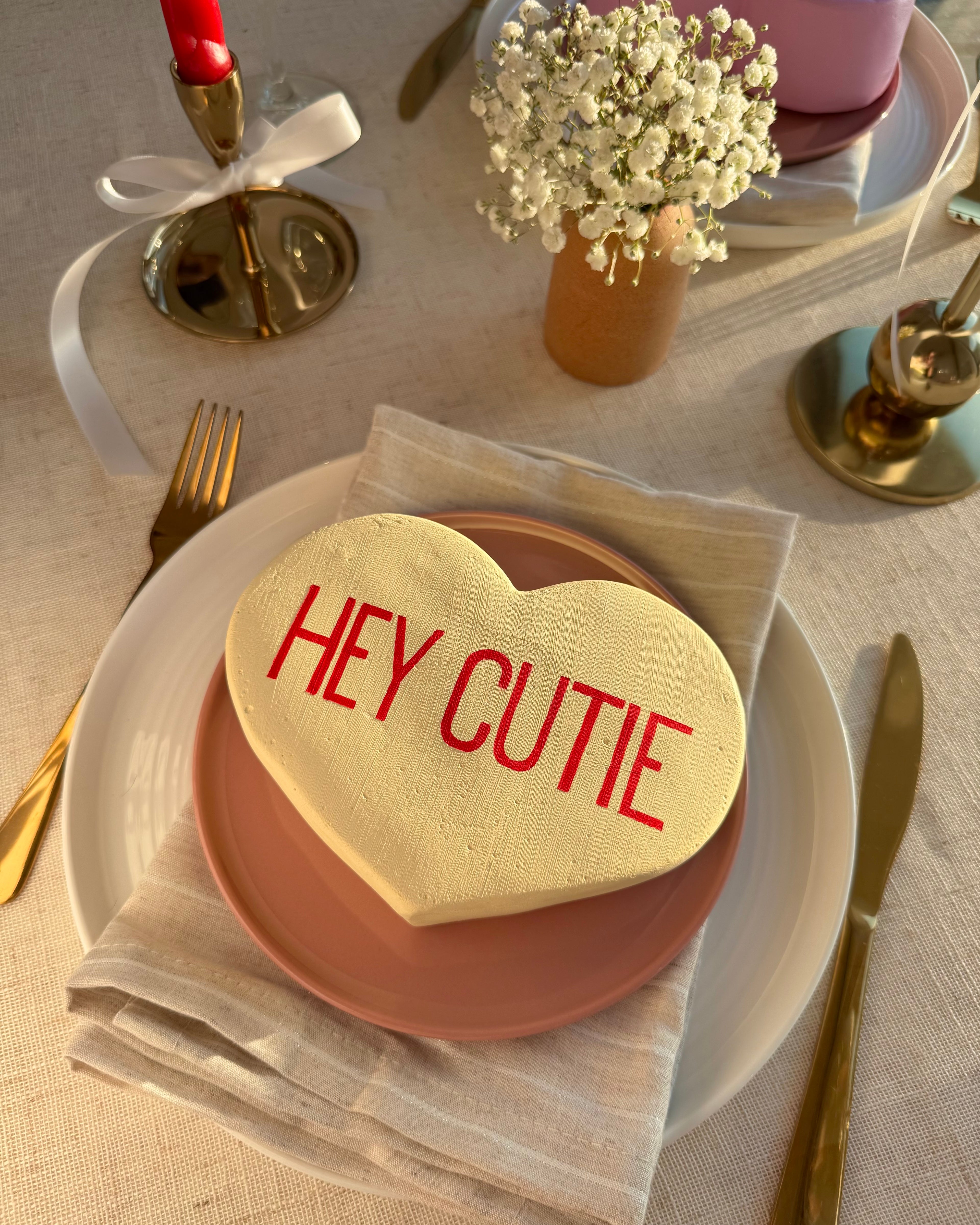 Yellow heart-shaped sculpture with 'HEY CUTIE' on a table setting with cutlery and flowers.