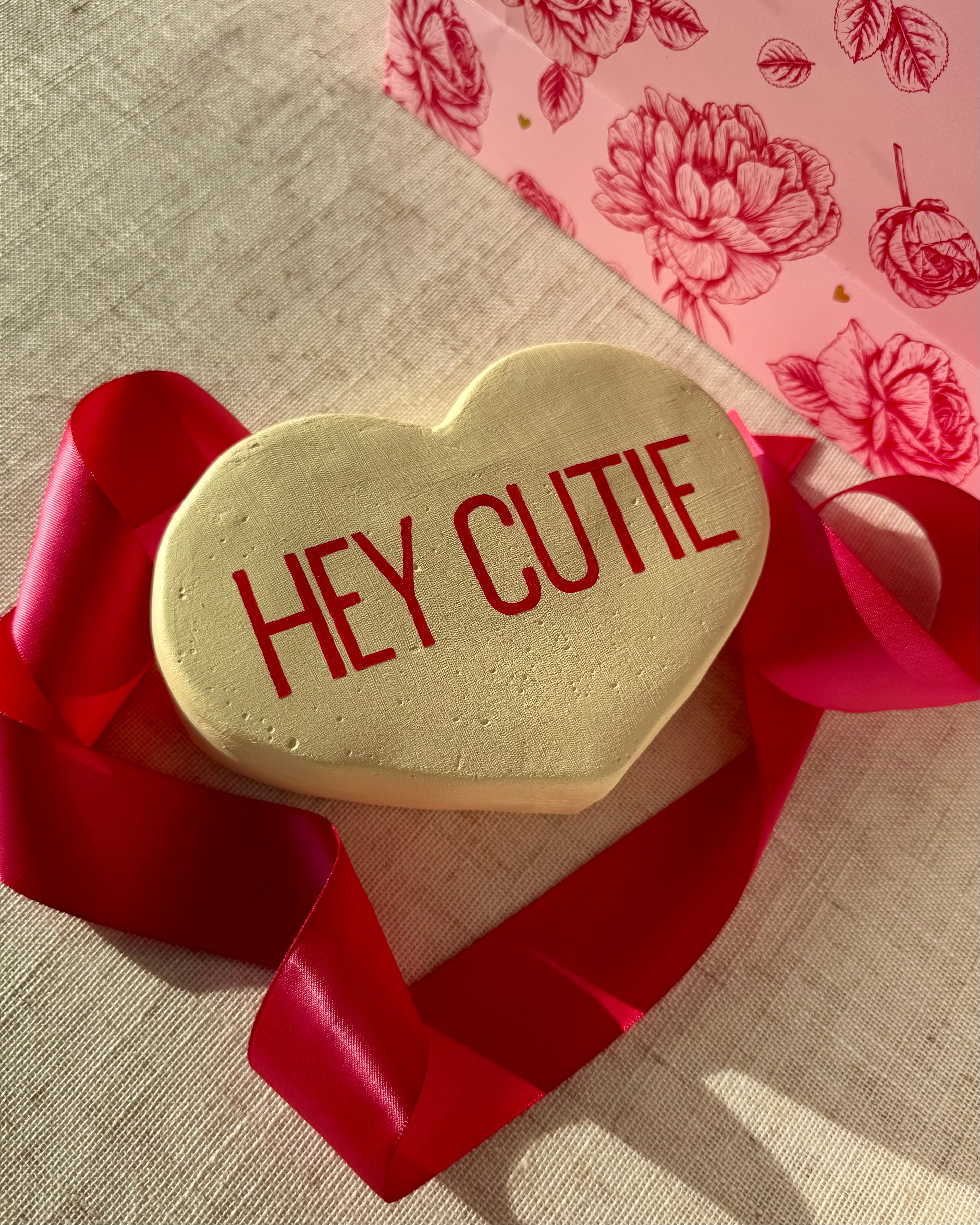 Yellow heart-shaped sculpture with 'HEY CUTIE' text on a red ribbon, against a floral-patterned background.