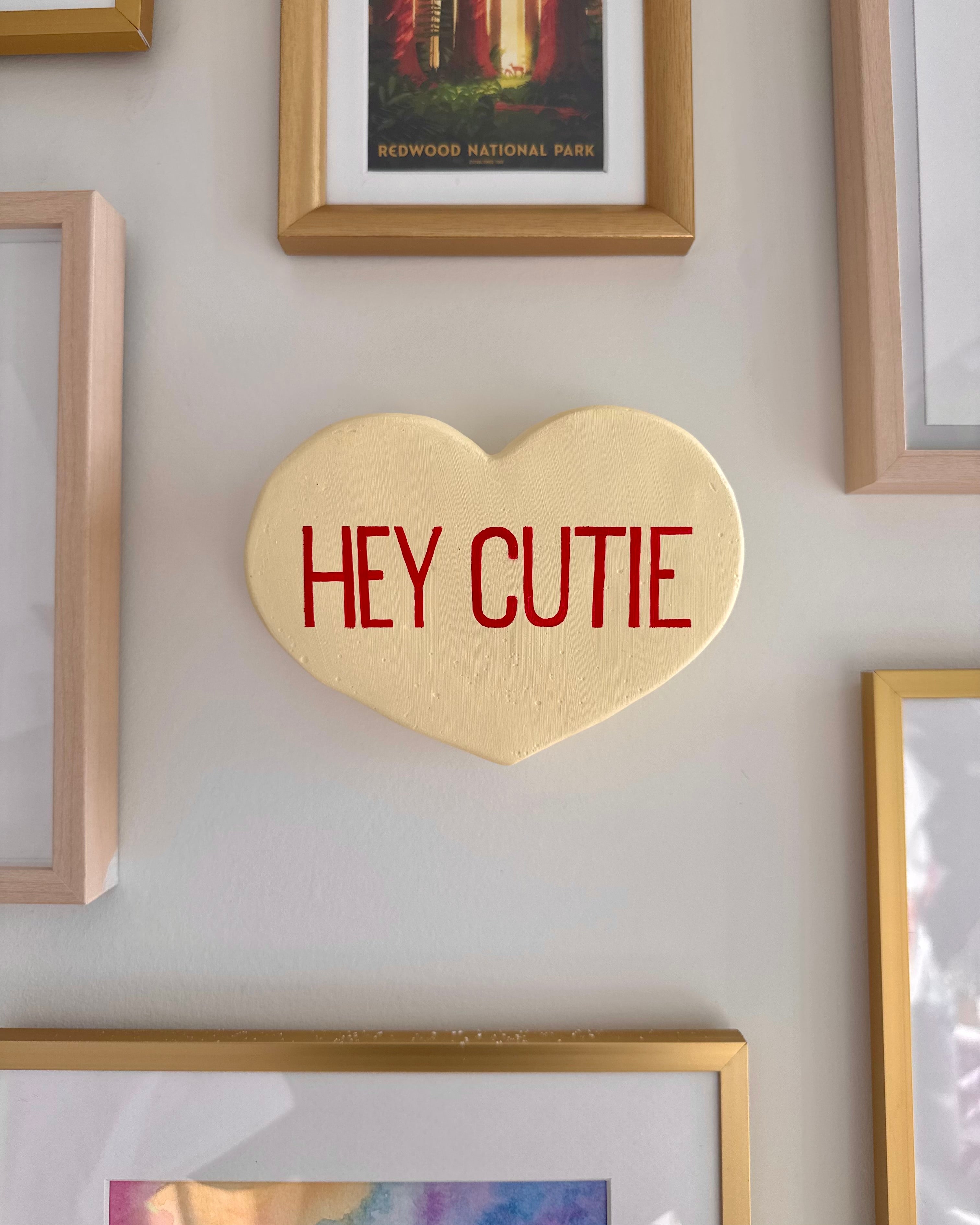 Yellow heart-shaped scultpure with 'HEY CUTIE' text on a wall with framed pictures