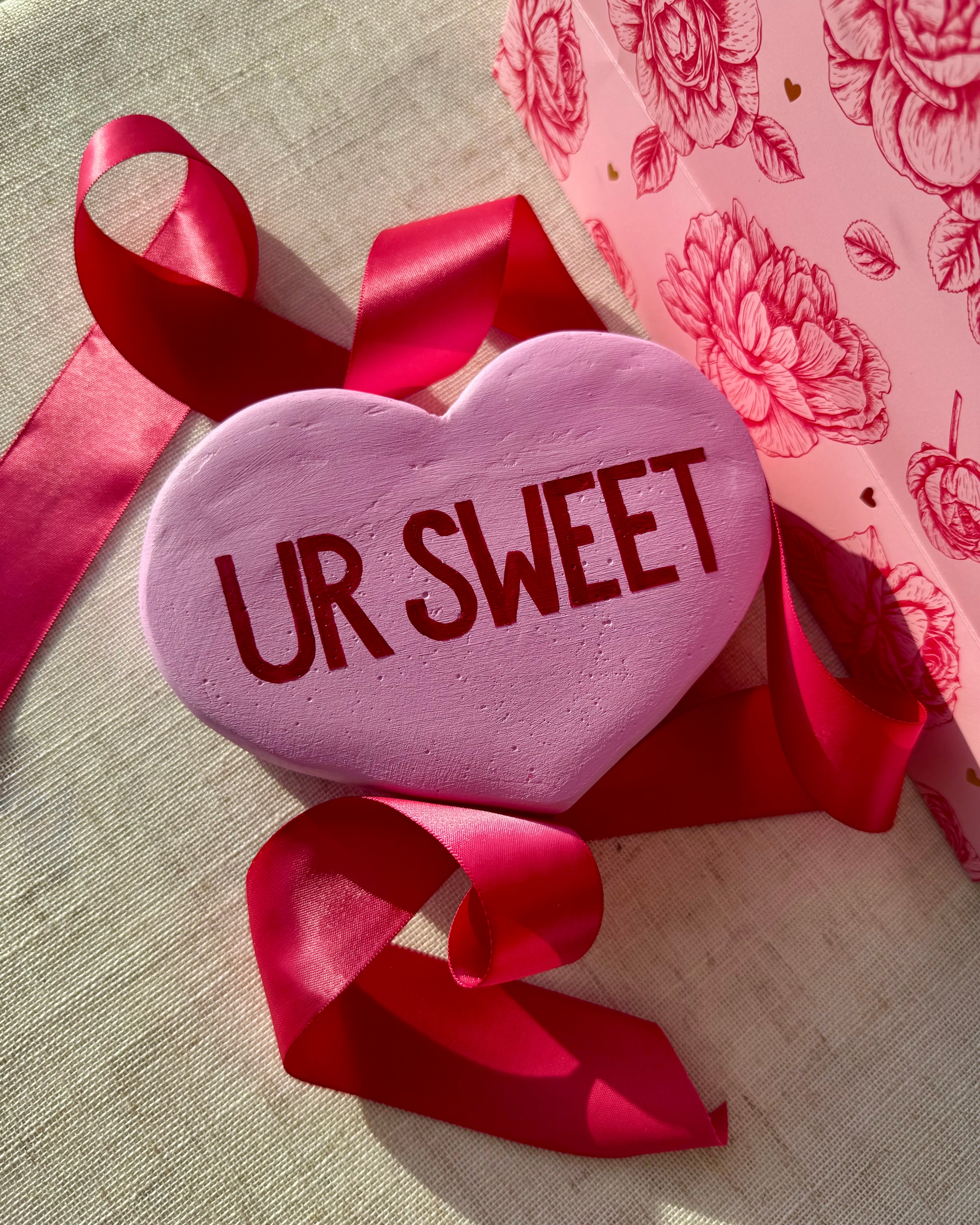 Light purple heart-shaped sculpture with 'UR SWEET' text, surrounded by red ribbons on a floral-patterned background.