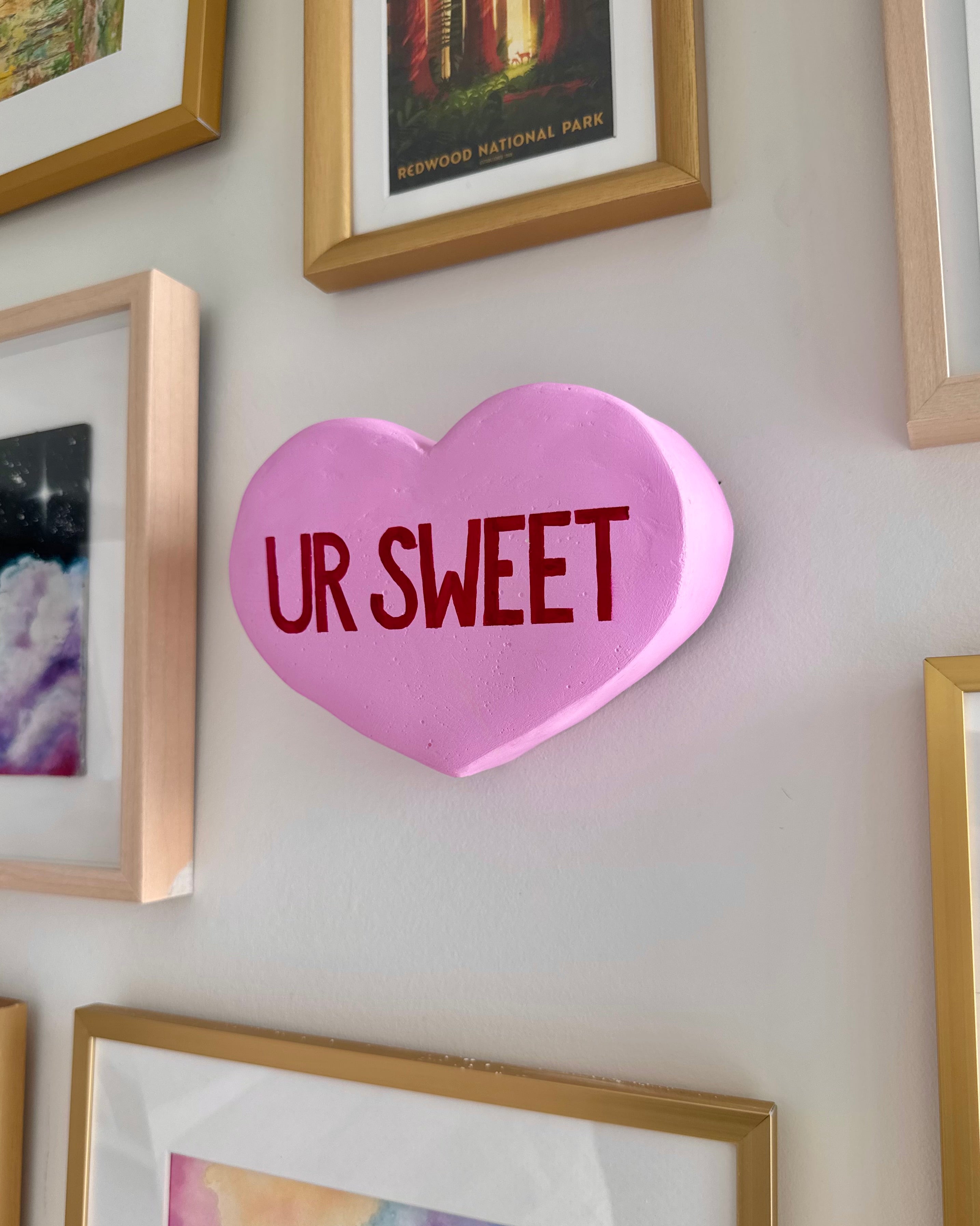 Light purple heart-shaped sculpture with 'UR SWEET' text on a wall with framed pictures.
