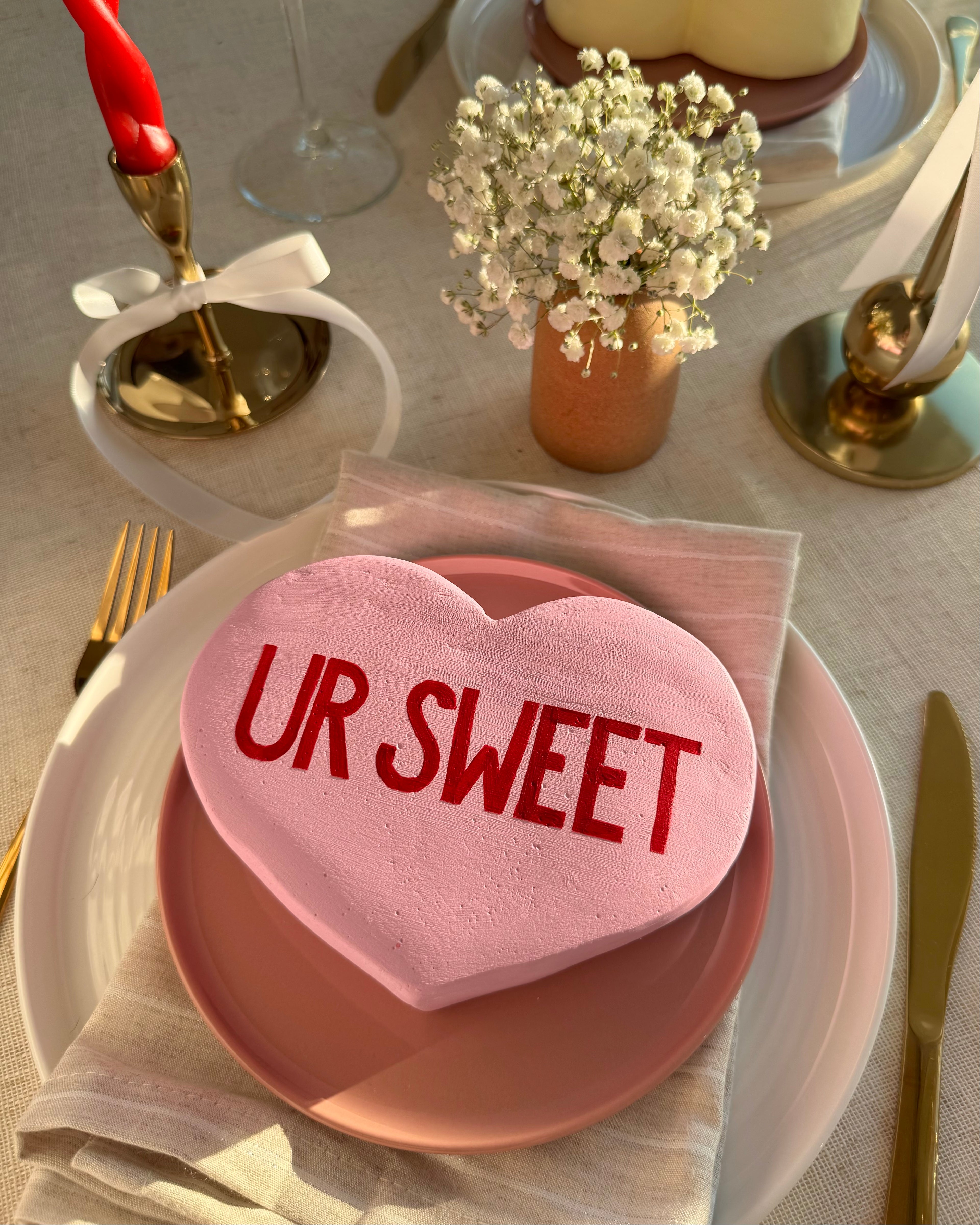Light purple heart-shaped sculpture with 'UR SWEET' text on a table setting with white flowers and candles.