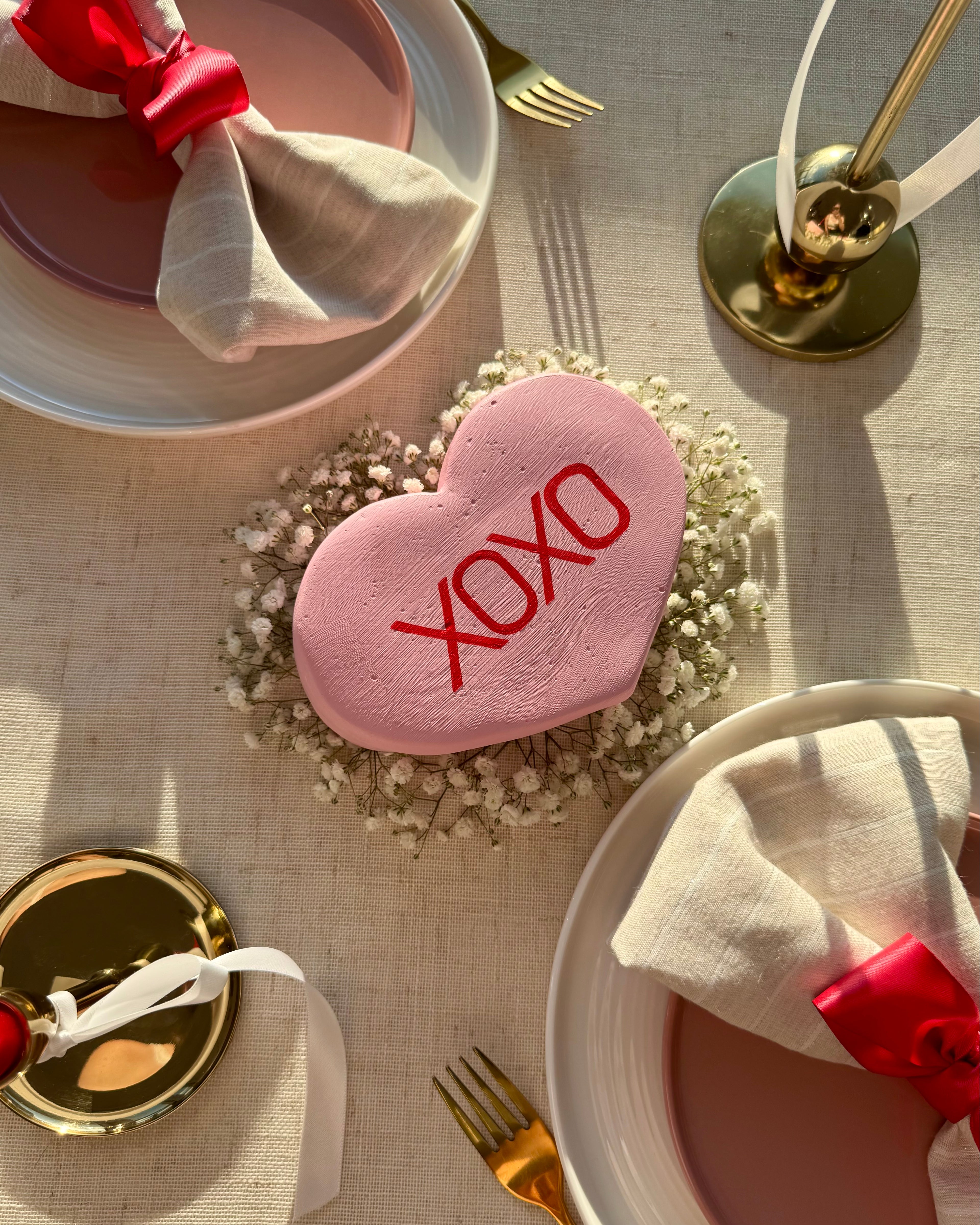 Pink heart-shaped candy sculpture with 'XOXO' on a table setting with plates, napkins, and cutlery.