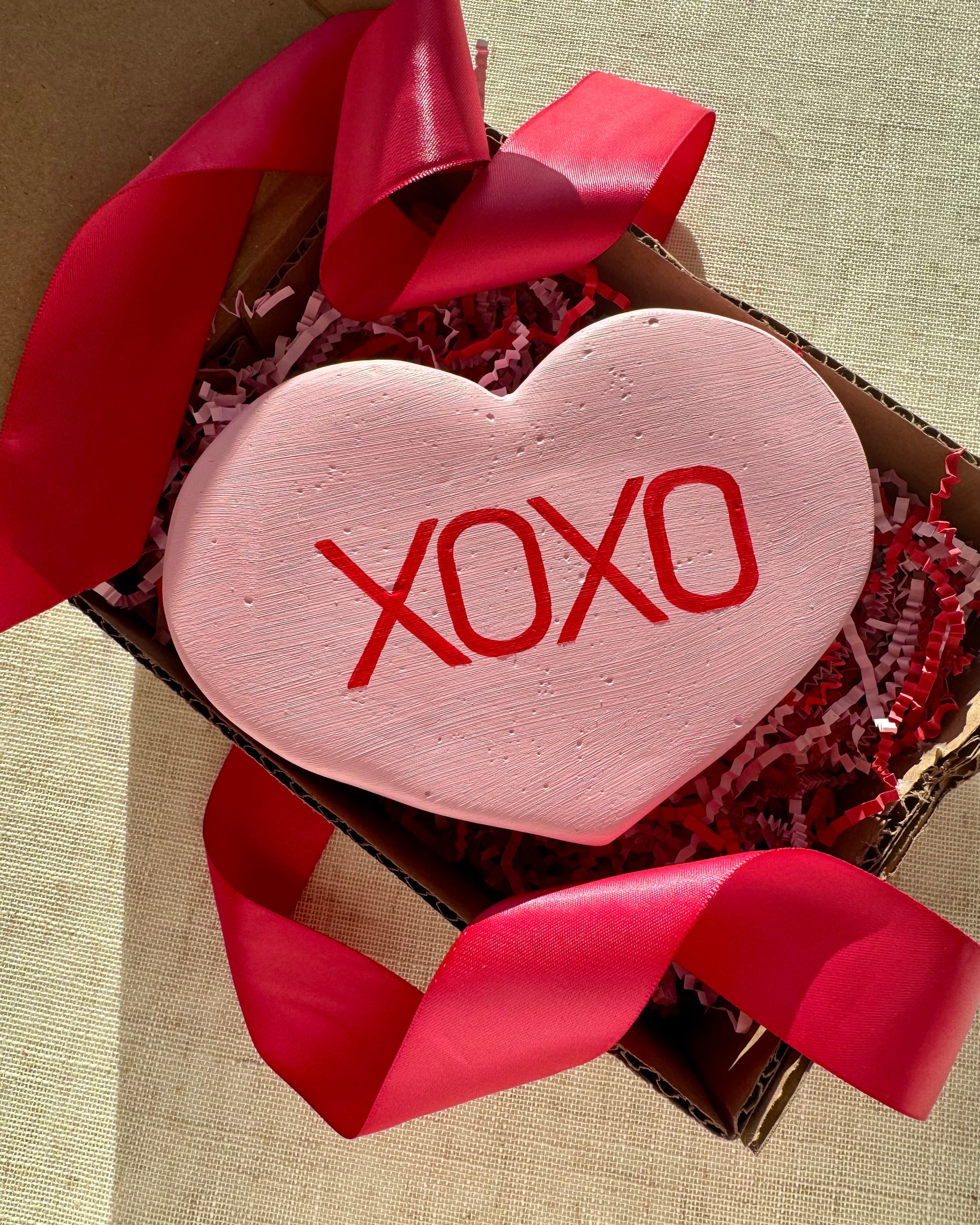 Pink heart-shaped sculpture with 'XOXO' text in a box with red ribbons on a beige background
